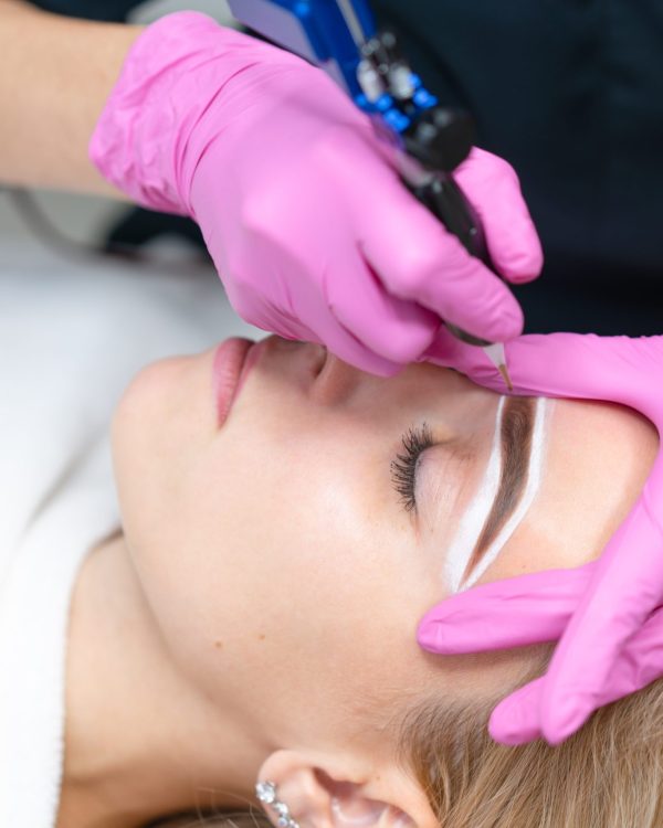 Cosmetology cabinet client lies on couch. Beautician applies permanent ink on eyebrows. Procedure of permanent eyebrow makeup. Close up. Beauty industry Pink gloves